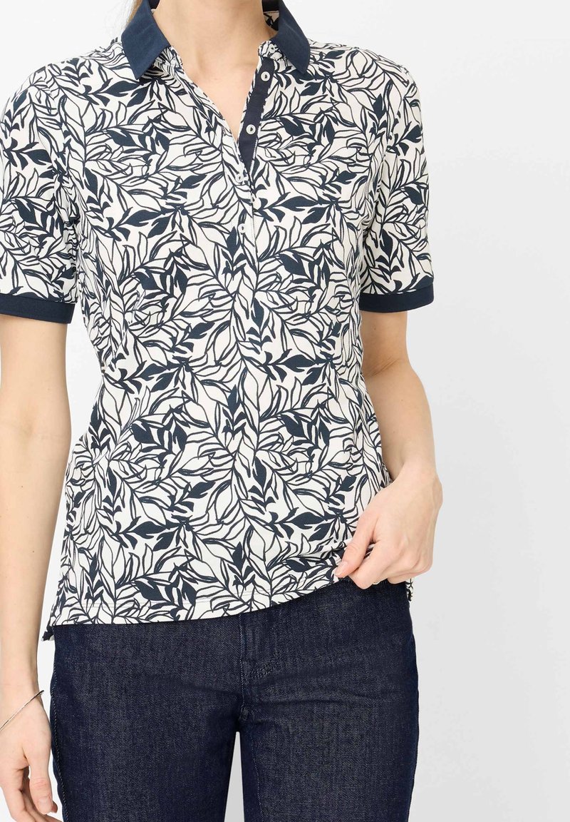 Femme portant un polo à manches courtes avec des motifs de feuilles bleu marine et un col, associé à un jean en denim bleu foncé.