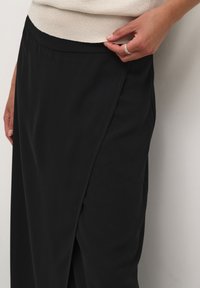 Jupe à enveloppe noire en tissu lisse, dotée d'une fente latérale et d'une taille ajustée. Main posée sur la taille pour un ajustement.