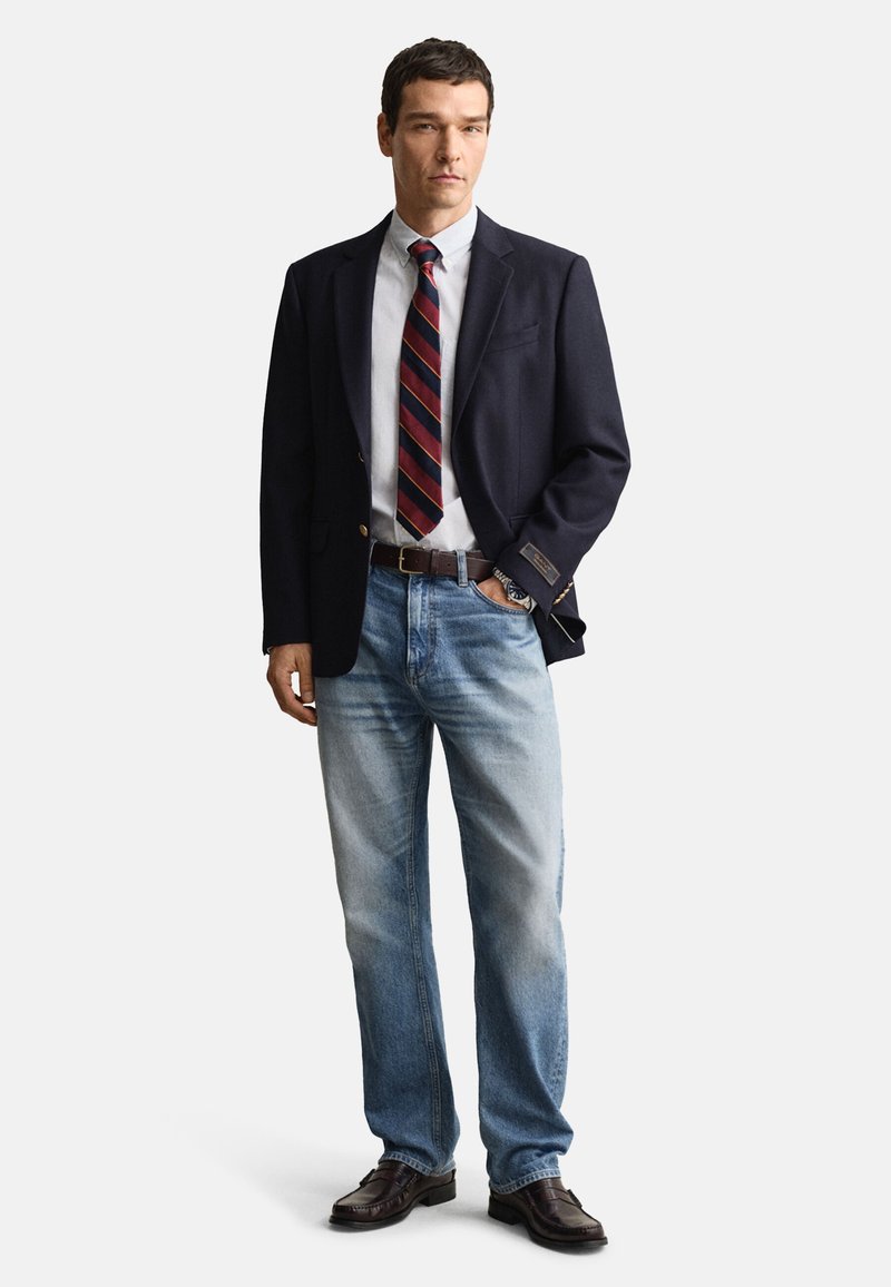 Blazer azul-marinho sobre uma camisa branca de vestir com uma gravata às riscas vermelha e preta. Jeans azul-claro e sapatos de couro castanhos completam o conjunto.