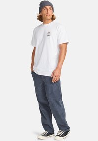 White cotton t-shirt with a circular logo, paired with dark blue pants and black sneakers. Styled with a gray knit beanie. Casual fit.