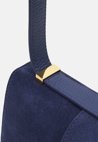 Borsa a tracolla in suede blu navy con una tracolla in cuoio testurizzato e un accento in metallo dorato al connettore. Design semplice e moderno.