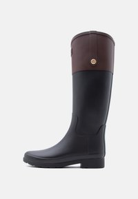 Ψηλή μαύρη λαστιχένια μπότα βροχής με σκουρό καφέ δερμάτινη μανσέτα και στρογγυλό χρυσό έμβλημα σε λευκό φόντο.