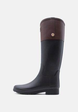 Hoge zwarte rubberen regenlaars met donkerbruine leren boord en ronde gouden embleem op een witte achtergrond.