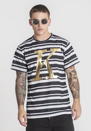 Młody mężczyzna o krótkich włosach, w okrągłych okularach przeciwsłonecznych, czarno-białej T-shircie w paski z dużą, złotą literą K oraz z tatuażami na ramionach.