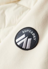 Toppa rotonda nera con testo "SUPERDRY" bianco e design geometrico cucita su tessuto in pile beige.