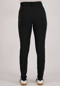 Zwarte fitted pantalon met een gladde textuur, twee achterzakken en riemlussen, gecombineerd met witte sneakers met donkere zolen.