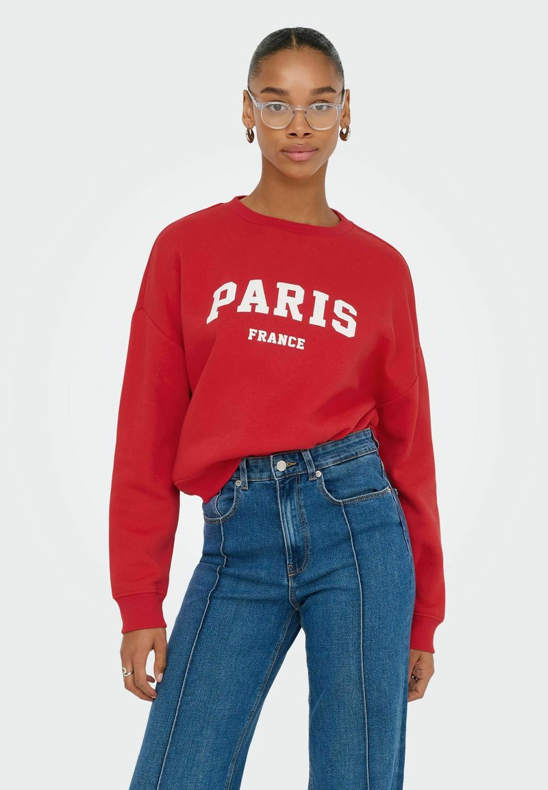 Rode sweatshirt met "PARIJS" en "FRANKRIJK" in witte letters, gecombineerd met high-waisted blauwe denim jeans. Duidelijke brilmonturen gedragen.