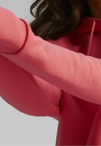 Felpa rosa con cappuccio, featuring polsini a coste e una texture di tessuto liscia. Primo piano sulla manica, evidenziando il colore e i dettagli del design.