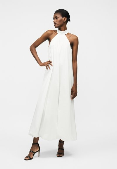 Femme portant une robe longue blanche sans manches à col licou et des talons hauts noirs à brides, debout avec une main sur la hanche.