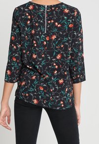 Schwarze Bluse mit floralem Muster und Dreiviertelärmeln, die rote und grüne Muster aufweist, einen runden Ausschnitt hat und einen Reißverschlussdetail auf der Rückseite.