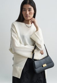 Suéter oversized em off-white com bainha canelada, combinado com uma mala de couro preta com detalhes em dourado e uma saia preta texturizada.