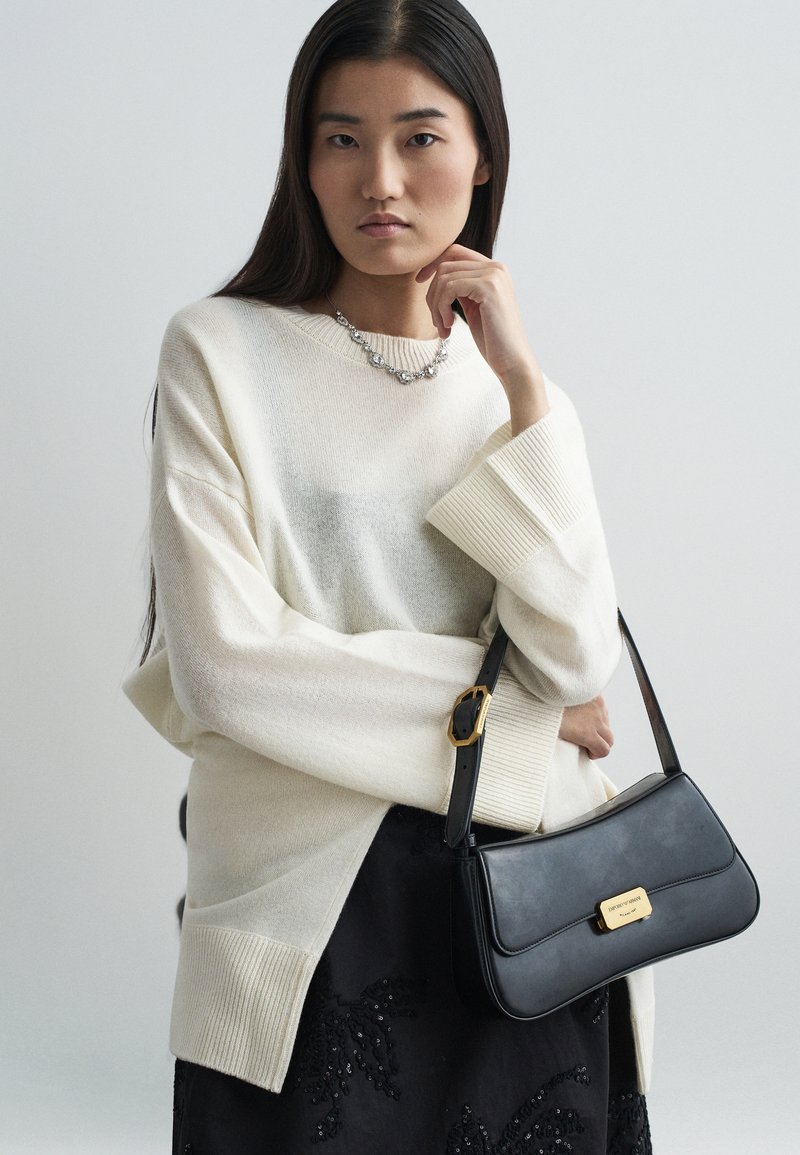 Suéter oversized em off-white com bainha canelada, combinado com uma mala de couro preta com detalhes em dourado e uma saia preta texturizada.