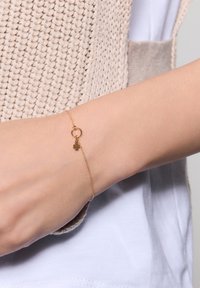 Złoty łańcuszek bransoletki z małym pierścionkiem i zawieszką w kształcie koniczyny. Delikatny design, gładka faktura i drobne detale w minimalistycznej estetyce.