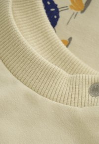 Close-up van de kraag van een romig gebreid sweater met ribbeltextuur, een decoratieve zilveren klavertje vier knop en een deel van een geprinte kattengezichtontwerp.