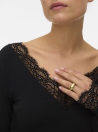 Žena nosi gornji dio s dugim rukavima od crne čipke, a na lijevoj ruci nosi zlatni prsten smješten blizu ključne kosti.