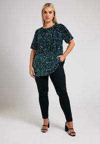 Top oversized di paillettes turchesi con maniche corte e orlo arrotondato, abbinato a pantaloni neri aderenti e sandali neri con tacco.