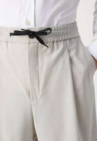 Pantaloni tailored grigi chiari con una texture morbida, dotati di un laccio nero e chiusura con bottone, e tasche laterali.