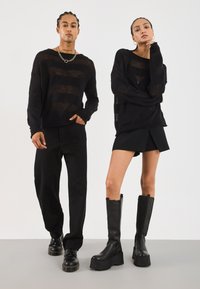 Schwarze Strickpullover mit gestreiftem Design, schwarze Hosen und einen geflämmten schwarzen Rock. Die Schuhe umfassen klobige schwarze Plateaustiefel.