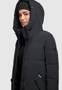 Chaqueta de plumas negra con un alto cuello y una capucha grande. Presenta un cierre de cremallera, botones a presión y un bolsillo lateral. Textura suave y diseño acolchado.