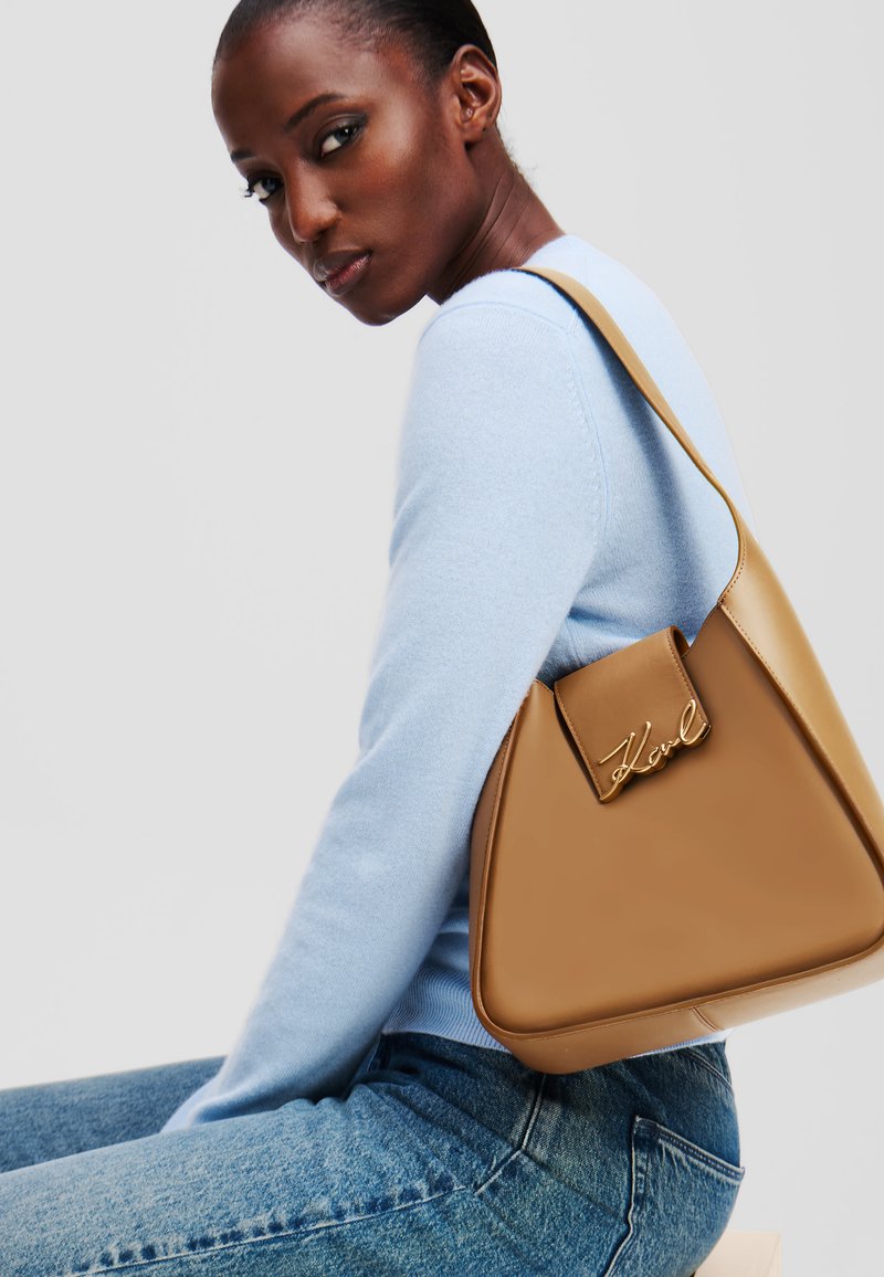 Tan triangular handbag with a smooth leather finish, featuring a flap closure with gold signature hardware. Worn on a shoulder.