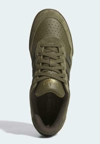 Zapatilla deportiva verde con parte superior texturizada y perforaciones, cordones y detalle del logo dorado en la lengüeta. Suela de goma y cuello acolchado.