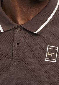 Brun polotröja med ett texturerat, andningsbart tyg. Har en krage med vita ränder, tre knappar och en liten Nike-logotyp i guld på en lapp.