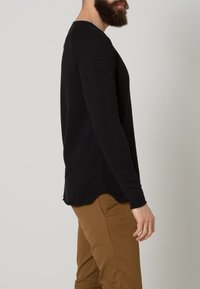 Svart långärmad skjorta i mjukt material, med en rundad nederkant och slim fit, kombinerad med beige byxor.