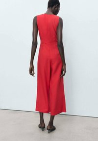 Red jumpsuit with a fitted bodice, wide legs, and a back zipper. Made of smooth fabric, features minimalist design and no visible patterns.