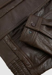 Materiale in pelle marrone scuro con superficie texturizzata, con cuciture visibili e bottoni a pressione, disposto in sezioni sovrapposte.