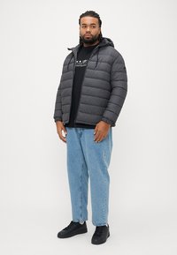 Γκρι μπουφάν puffer με κουκούλα, με καπιτονέ σχεδιασμό. Φοράει πάνω από μαύρο πουκάμισο, συνδυασμένο με ανοιχτό μπλε τζιν και μαύρα αθλητικά παπούτσια.