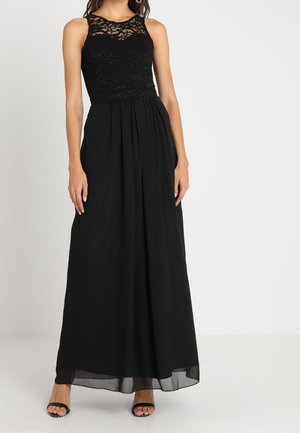 Vrouw draagt een mouwloze zwarte kanten topjurk met een vloeiende chiffon rok en zwarte open-toe hakken tegen een effen achtergrond.