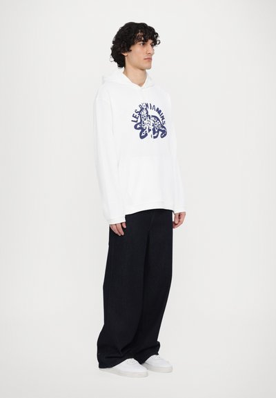 Sweat à capuche blanc avec une poche frontale, orné d'un design graphique bleu. Associé à un pantalon en denim foncé ample et des baskets blanches.