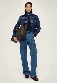 Námořnická péřová bunda se zipem, kombinovaná se světle modrými cargo džíny s postranními kapsami. Model drží černou tašku tote.