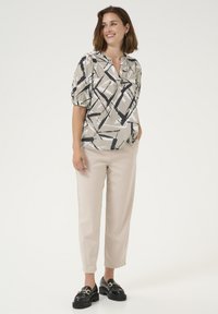 Lichte, kortärmelige blouse in beige met zwarte abstracte patronen; gecombineerd met lichtbeige broeken en zwarte loafers.