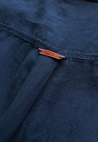 Tela azul oscuro con un pliegue cosido y una pequeña etiqueta naranja cosida que dice "SUPERDRY" centrada en la costura.