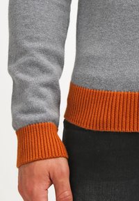 Grau gestrickter Pullover mit gerippten orangen Bündchen und Saum. Der Stoff hat eine glatte Textur mit einem lässigen Schnitt, der kontrastierende Farben betont.