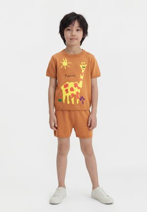 Παιδί με σκούρα μαλλιά στέκεται φορώντας πορτοκαλί μπλουζάκι με κοντά μανίκια και σορτς, που έχουν σχέδιο με κίτρινη και πορτοκαλί καμηλοπάρδαλη και ήλιο, καθώς και λευκά αθλητικά παπούτσια.