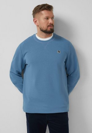 Man met baard draagt een blauw sweatervest met ronde hals over een wit shirt, staand tegen een lichtgrijze muur, handen achter de rug.