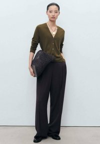 Olive green cardigan with five buttons, paired with dark wide-leg trousers. Holding a dark woven bag. Simple necklace accentuates the outfit.