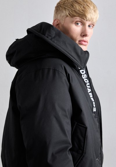 Black padded jacket with a large hood, featuring a zip closure, snap buttons, and a prominent logo on the front. Smooth texture.