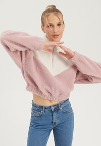 Pull en polaire rose clair et blanc, avec un col zippé mi-hauteur et un design court. Doté d'un tissu texturé et d'une taille élastiquée.