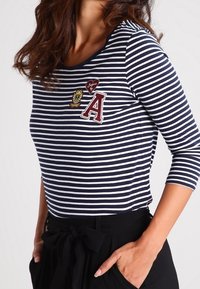 Marinblå och vit randig långärmad topp med rundad halsringning, med broderade lappar i olika färger på bröstet. Material i bomullsblandning.