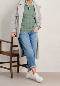 Trench-coat gris clair porté sur un pull vert en maille à motif texturé, associé à un jean bleu décontracté et des baskets blanches.