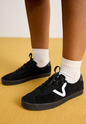 Personne portant des baskets noires avec un logo blanc et des chaussettes blanches debout sur un sol jaune.