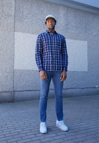 Tamsiai mėlynos, raudonos ir baltos spalvos langeliais skirta marškiniai su mygtukais; siauri mėlyni džinsai; baltos sportbačiai; šviesios spalvos kepurė; stovi prieš tekstūruotą sieną.