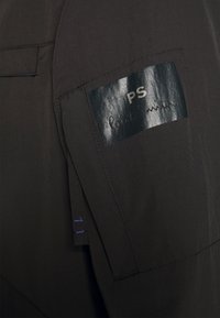 Manche en tissu foncé avec un patch rectangulaire noir brillant portant l'inscription "PS Paul Smith" cousu sur le haut du bras.