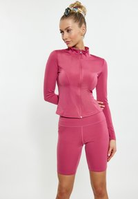 Completo sportivo aderente in rosa; maglione con zip a maniche lunghe e colletto alto abbinato a leggings corti. Tessuto liscio con elasticità per il massimo comfort.
