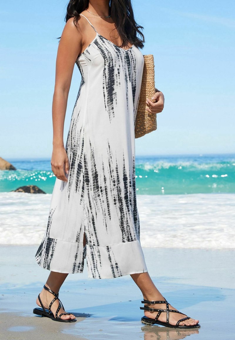 Vestido maxi con estampado blanco y negro, tirantes finos, aberturas laterales y un diseño fluido. Combinado con sandalias negras de tiras y un bolso de mimbre.
