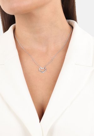 Collana d'argento con pendente a forma di cuore geometrico, caratterizzata da una catena fine e un design minimalista. Indossata sopra un blazer bianco.