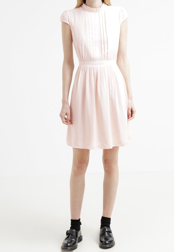 Femme portant une robe rose pâle à manches courtes avec des plis, des chaussettes noires et des chaussures noires à boucle, debout devant un fond blanc uni.